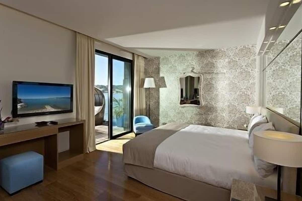 Habitación de hotel moderna con una cama grande, un televisor sobre un escritorio de madera, un balcón con vistas al mar y una pared de mármol.