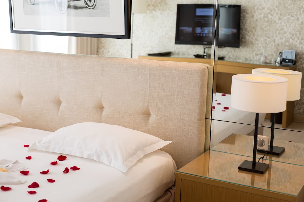Una habitación de hotel con cama, almohadas blancas, pétalos de rosa, una mesita de noche con lámpara, televisor en la pared y un mostrador de recepción.