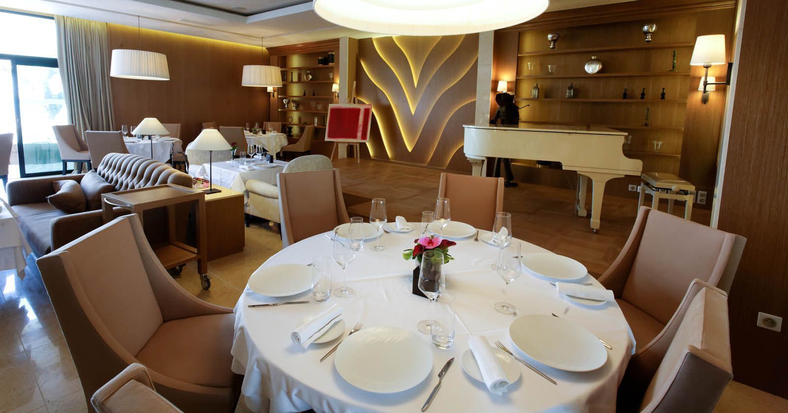 A dining area with round tables set for four, each with white tablecloths, plates, and cutlery. There is a piano in the background, and the room has a warm, modern decor with wooden paneling and a large, curved light fixture.