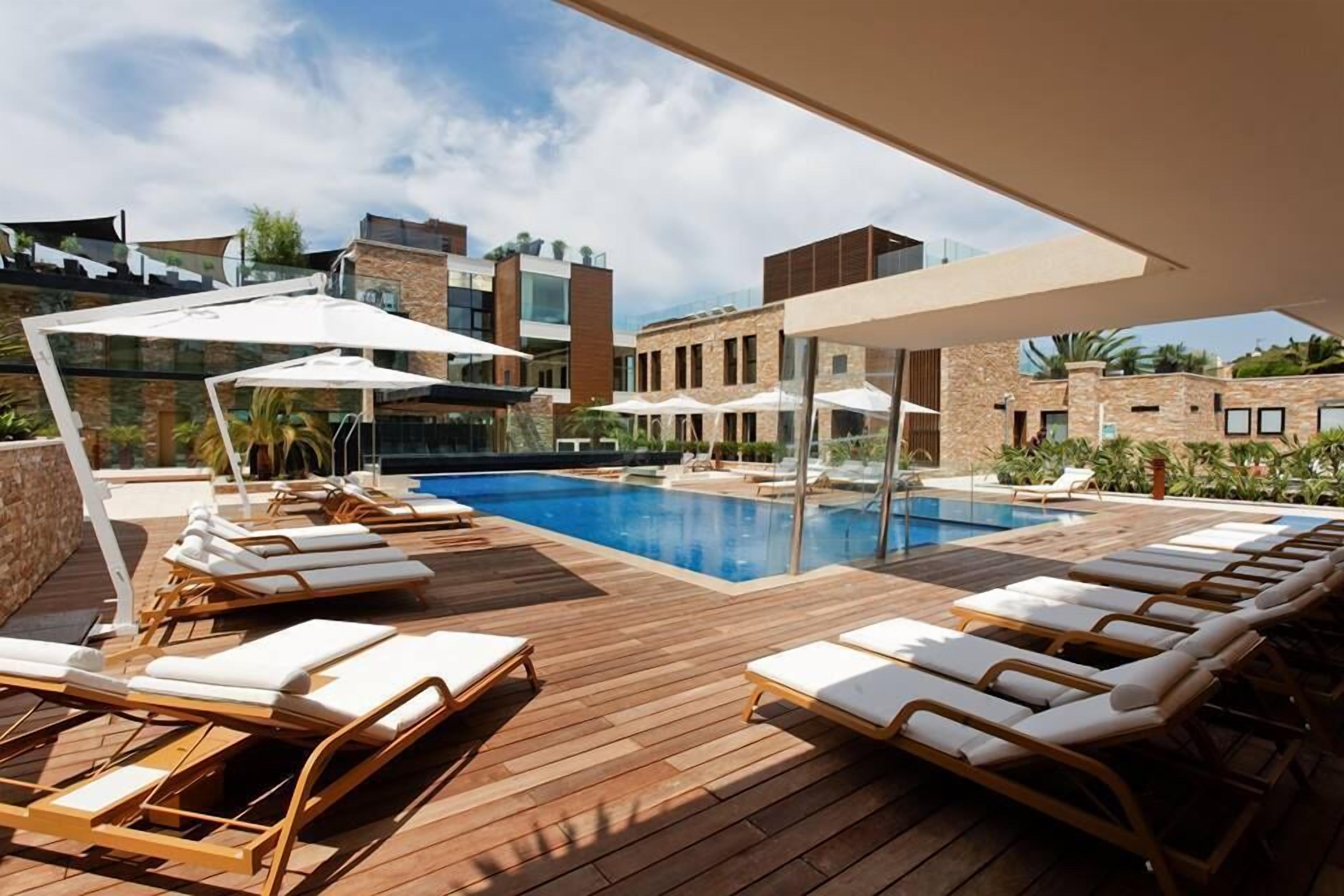 A poolside area with lounge chairs and umbrellas, surrounded by modern buildings with large windows.