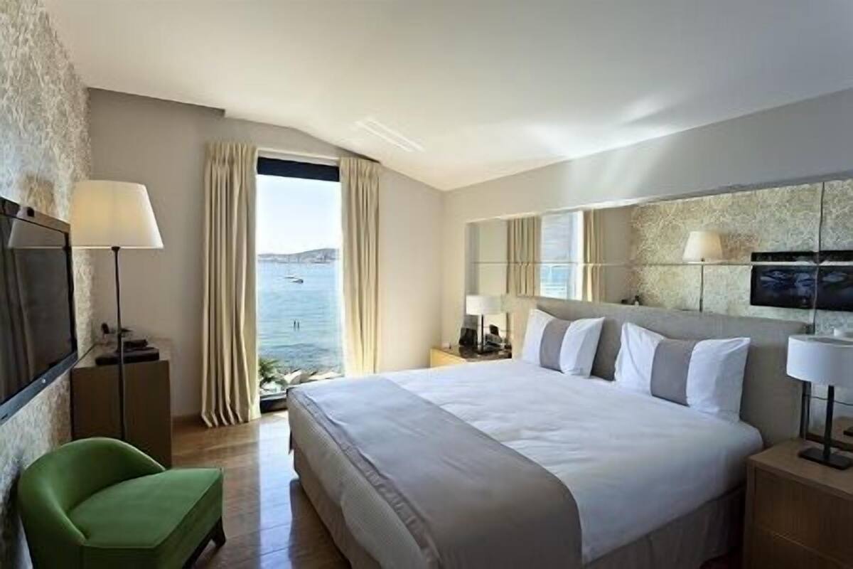 Habitación de hotel con una cama grande, una silla verde, una mesita de noche con lámpara y vistas al mar a través de una ventana.