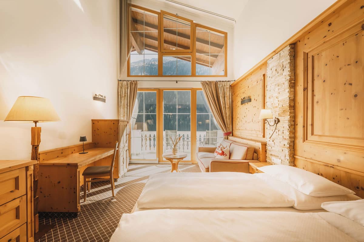 Ein Zimmer mit hohen Decken, einem Bett, Schreibtisch und Stuhl, großen Fenstern und einer Steinwand.