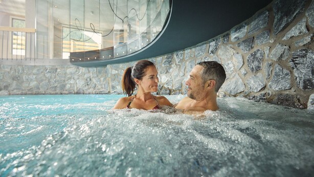 Un couple dans un jacuzzi, avec un mur en pierre et une paroi en verre en arrière-plan.
