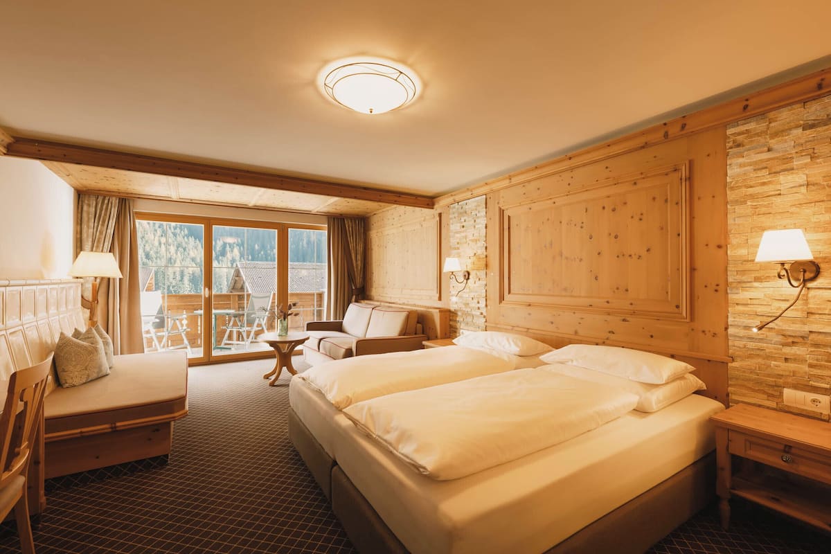 Ein Hotelzimmer mit zwei Betten, einem Schlafsofa, einem kleinen Tisch und einem Fenster mit Blick auf Bäume.
