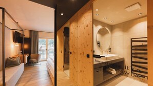 Une chambre d’hôtel moderne avec un plancher en bois, un canapé, un bureau, un grand miroir et une salle de bain équipée d’un porte-serviettes.