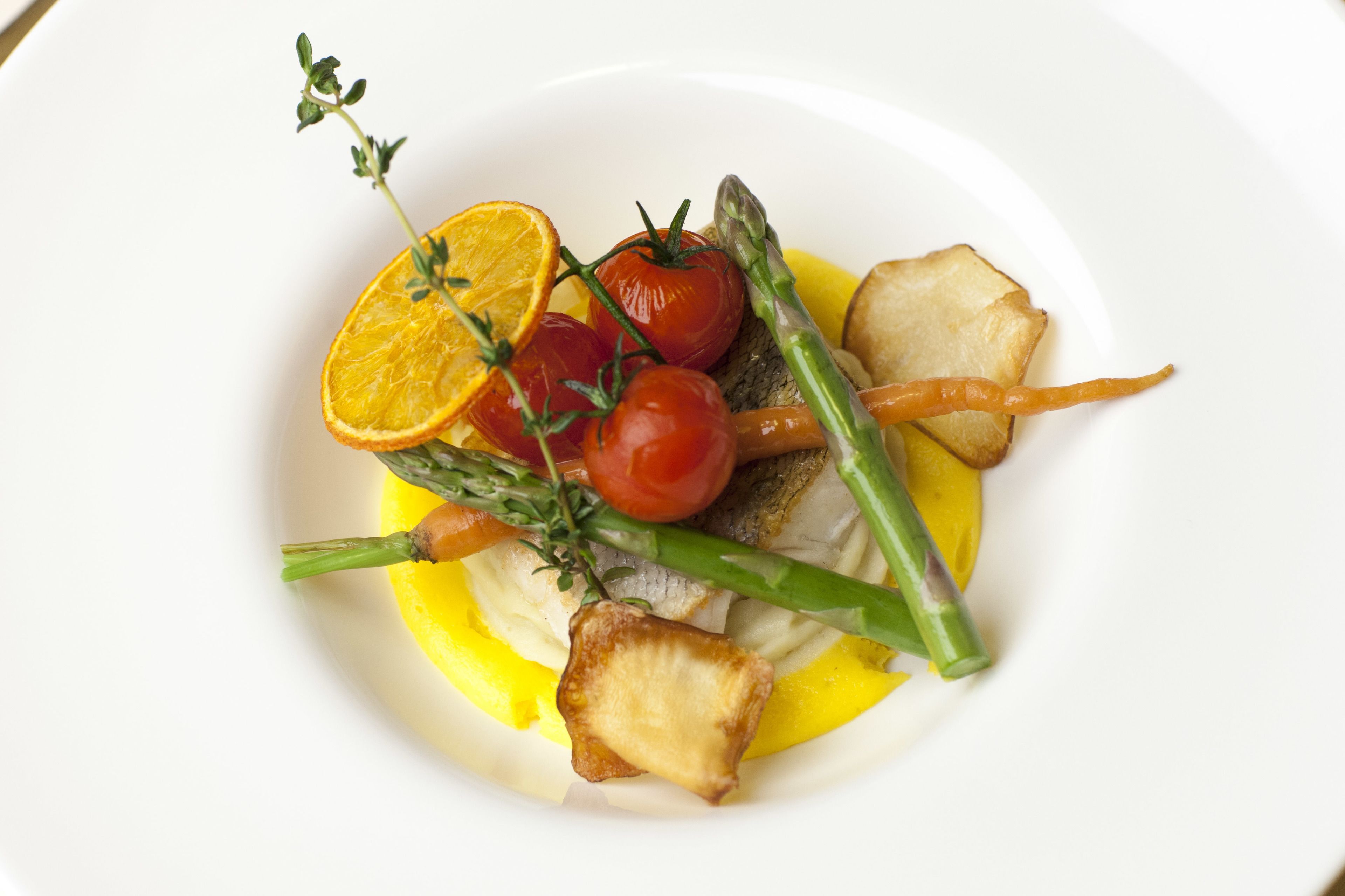 Ein weißer Teller mit Fischfilet, Cocktailtomaten, Spargel und einer Orangenscheibe.