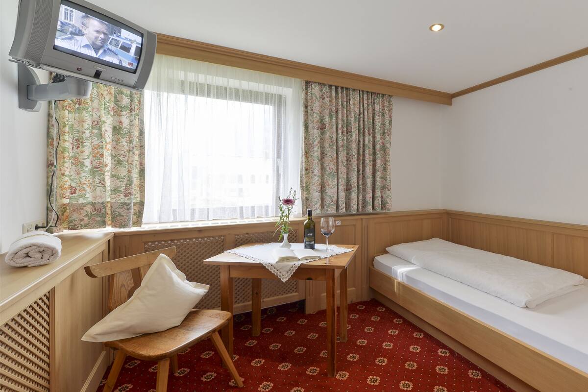 Une chambre d’hôtel de taille modeste, comprenant un lit, une petite table avec un livre, une chaise, un téléviseur et une fenêtre agrémentée de rideaux à motifs floraux.