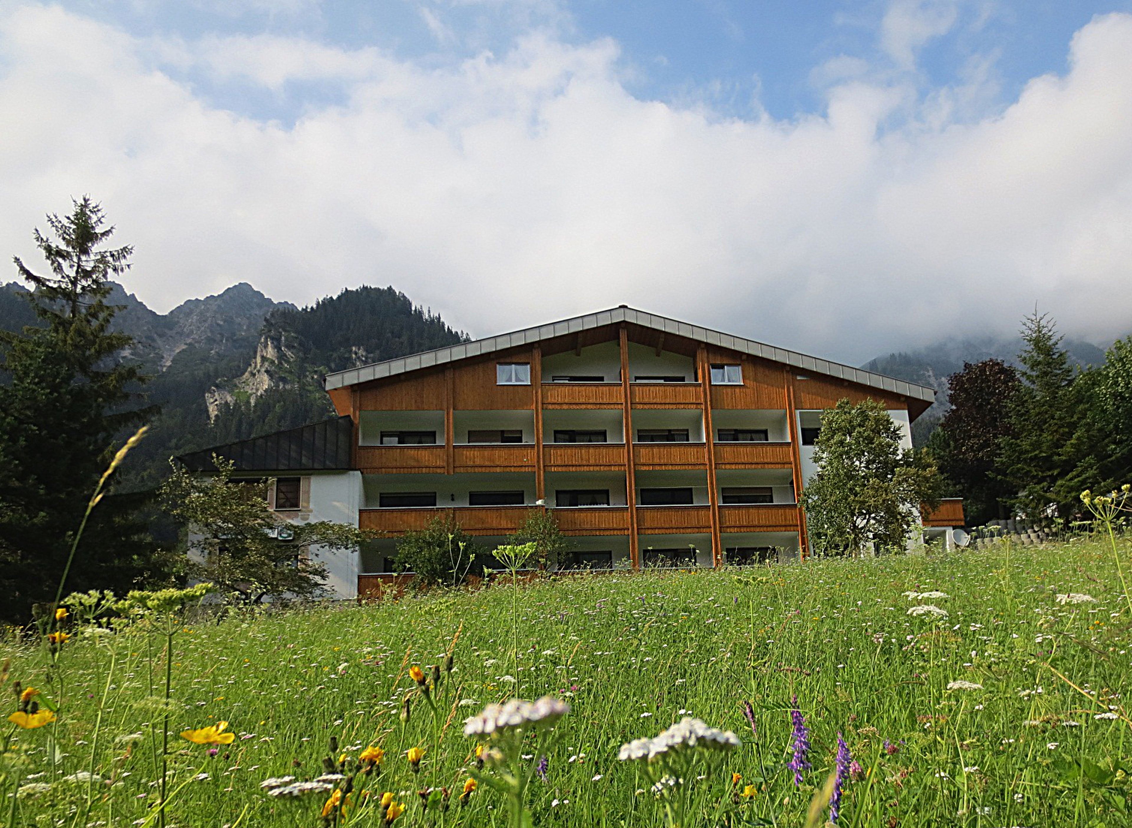 A multi-story building with a wooden facade, surrounded by a lush meadow and mountains in the background.