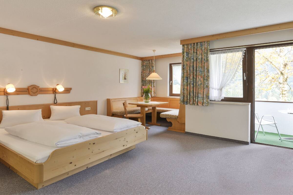 Une chambre d’hôtel avec un lit en bois, une petite table avec une chaise, une fenêtre avec des rideaux et une lampe.