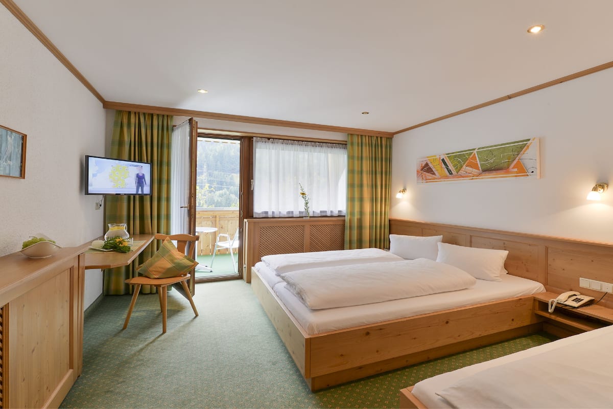 Une chambre d’hôtel avec un lit en bois, un bureau avec une chaise, une télévision et un balcon avec des rideaux.