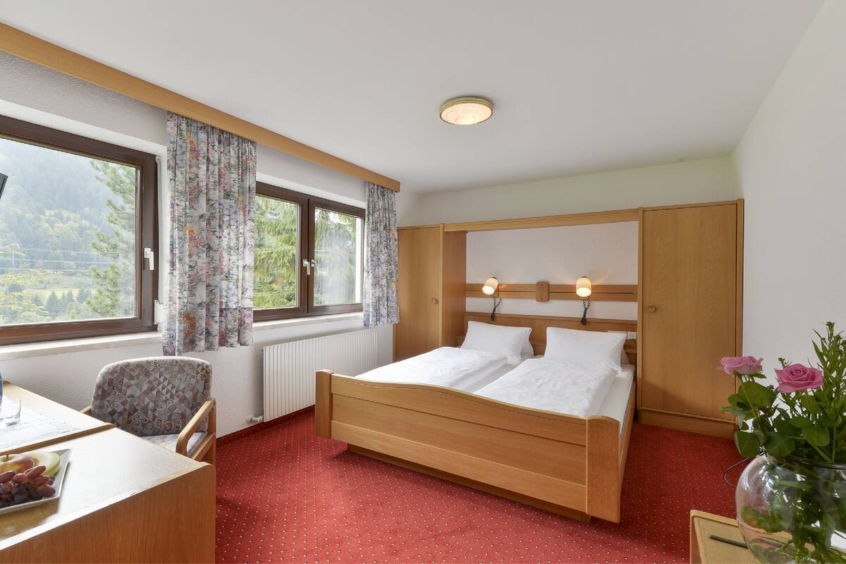 Une chambre d’hôtel avec un lit en bois, un bureau avec une chaise, une fenêtre avec des rideaux, un radiateur et un vase avec des fleurs.