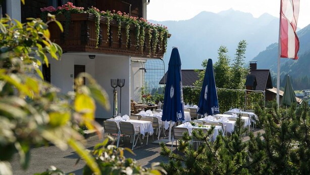 Ein Essbereich im Freien mit Tischen und blauen Sonnenschirmen, umgeben von Grünpflanzen und Blick auf eine Berglandschaft.