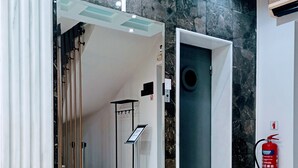 Un hall d’entrée moderne avec un sol en marbre, un ascenseur en verre, un extincteur et un présentoir d’information en métal.