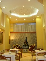 Uma sala de jantar com mesas e cadeiras, teto com iluminação embutida e uma janela grande com cortinas.