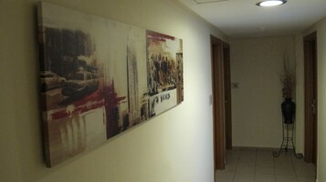 Um corredor com uma grande pintura abstrata na parede, uma porta com o número 74 e um vaso preto sobre um suporte no canto.
