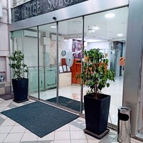 Entrada de hotel com portas de vidro, plantas em vasos, balcão de recepção e sinalização de saída visível.