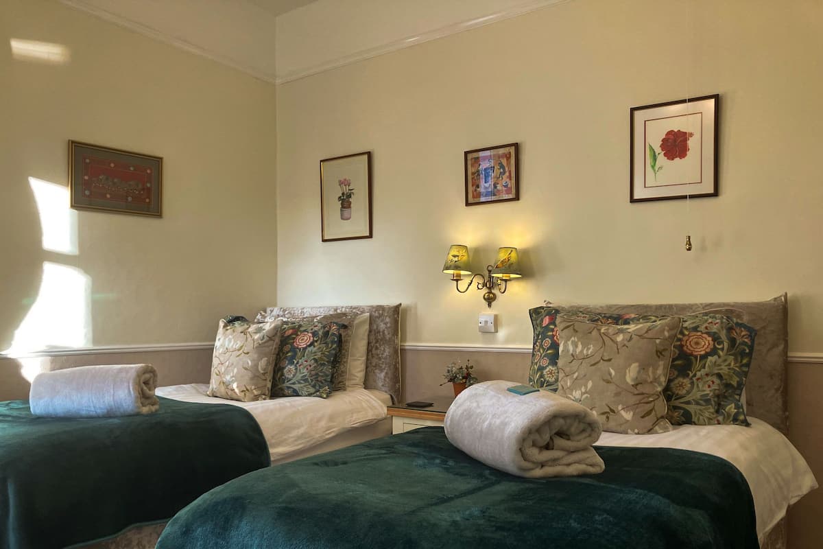 A hotel room with two beds, each with a green blanket and multiple pillows. There are wall-mounted lamps and framed pictures on the walls.