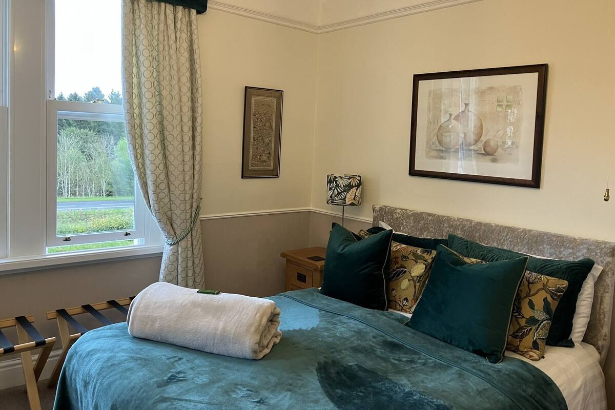 A neatly made bed with a blue cover, a window with a view of greenery, and a framed picture on the wall.