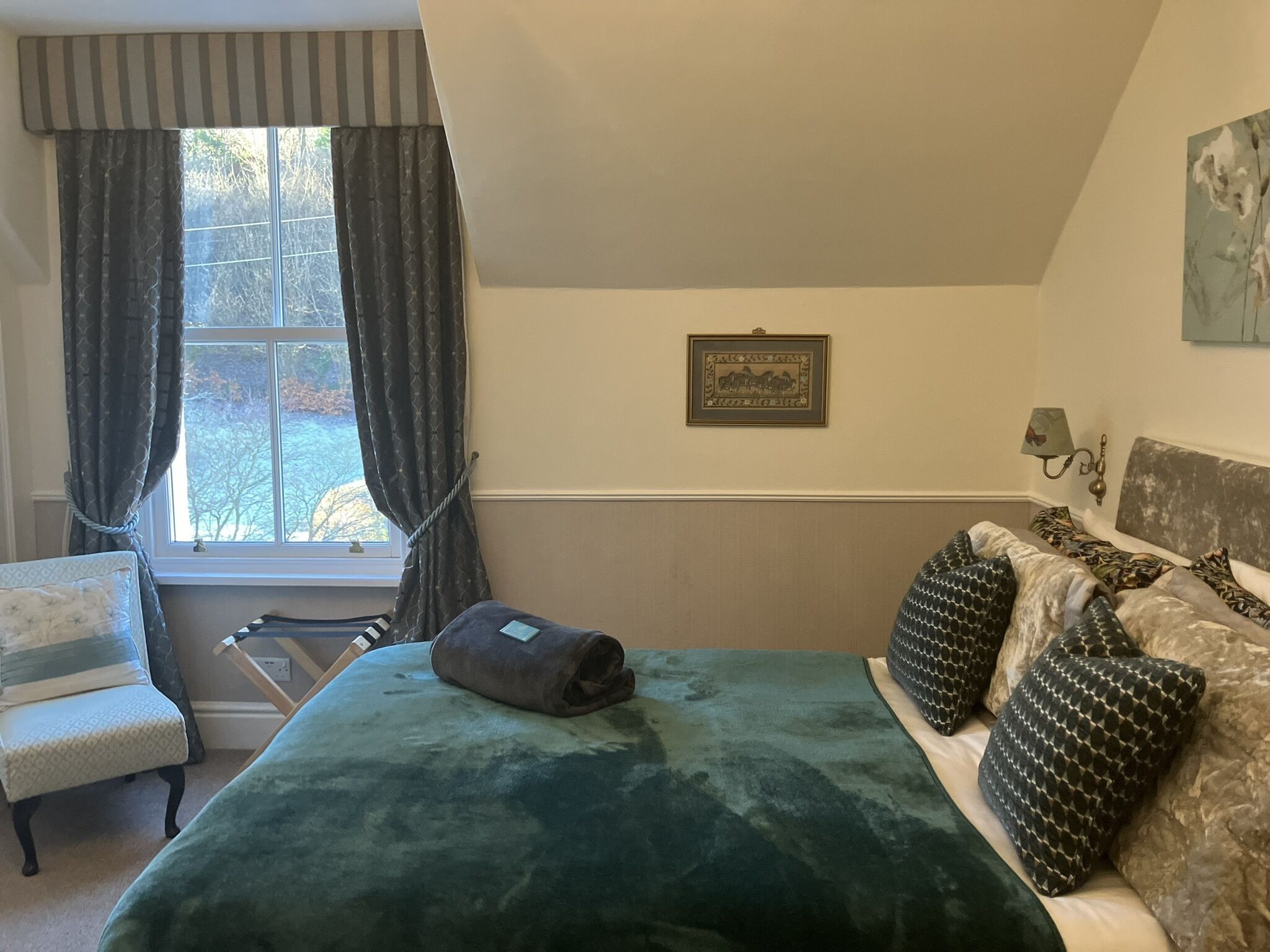 A bedroom with a bed, a chair, a window with curtains, a wall-mounted lamp, and a framed picture.