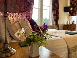 A neatly made bed with a white comforter and pillows, a bedside table with a lamp and a vase of flowers, and a window with curtains. - Maple Bank (Keswick)