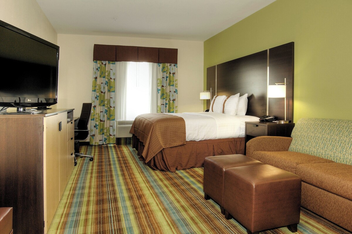 A hotel room with a bed, a TV, a desk, a chair, a sofa, and a patterned carpet.