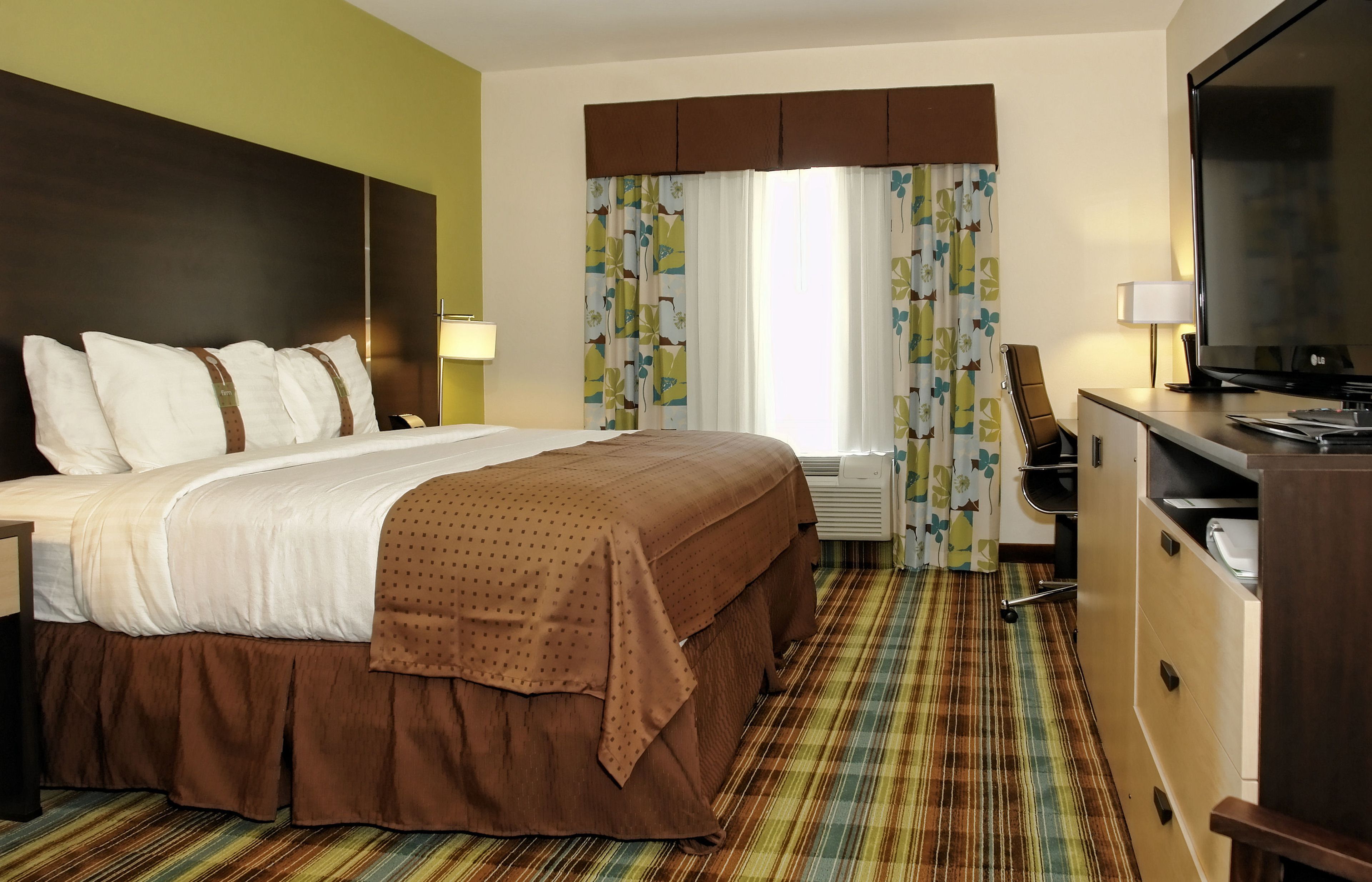 Ein Hotelzimmer mit einem großen Bett, einem Schreibtisch mit Computer, einem Fernseher und einem Fenster mit Vorhängen.