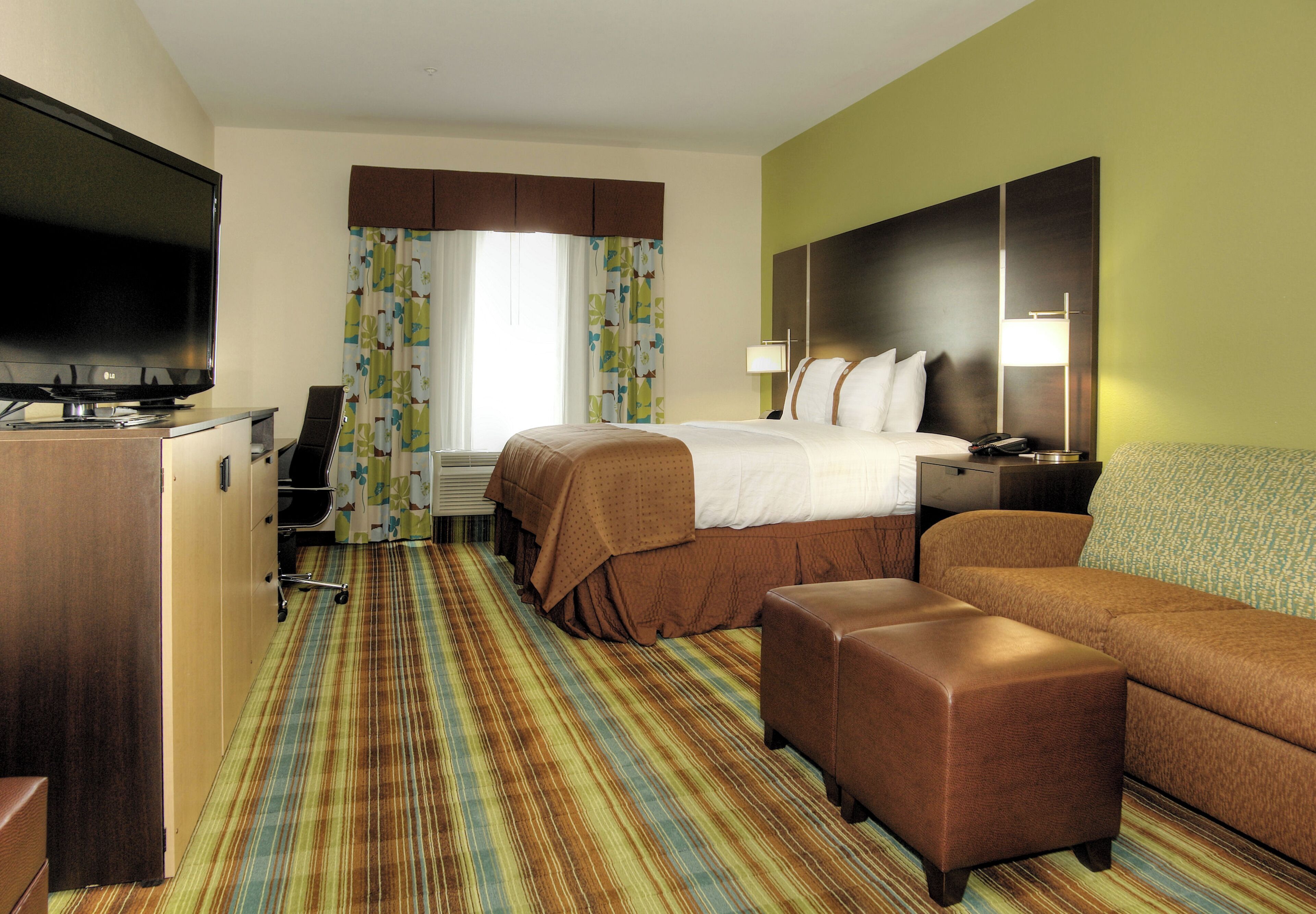 A hotel room with a bed, a TV, a desk, a chair, a sofa, and a patterned carpet.