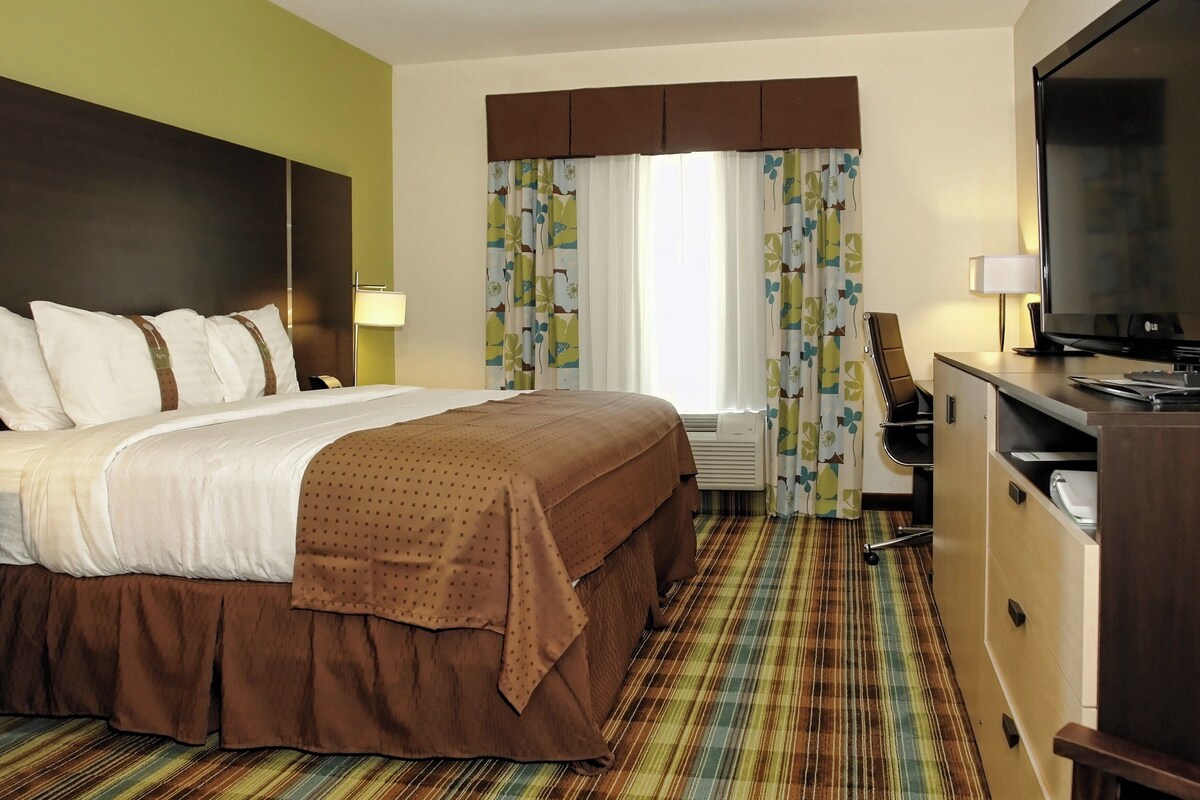 A hotel room with a large bed, a desk with a computer, a TV, and a window with curtains.