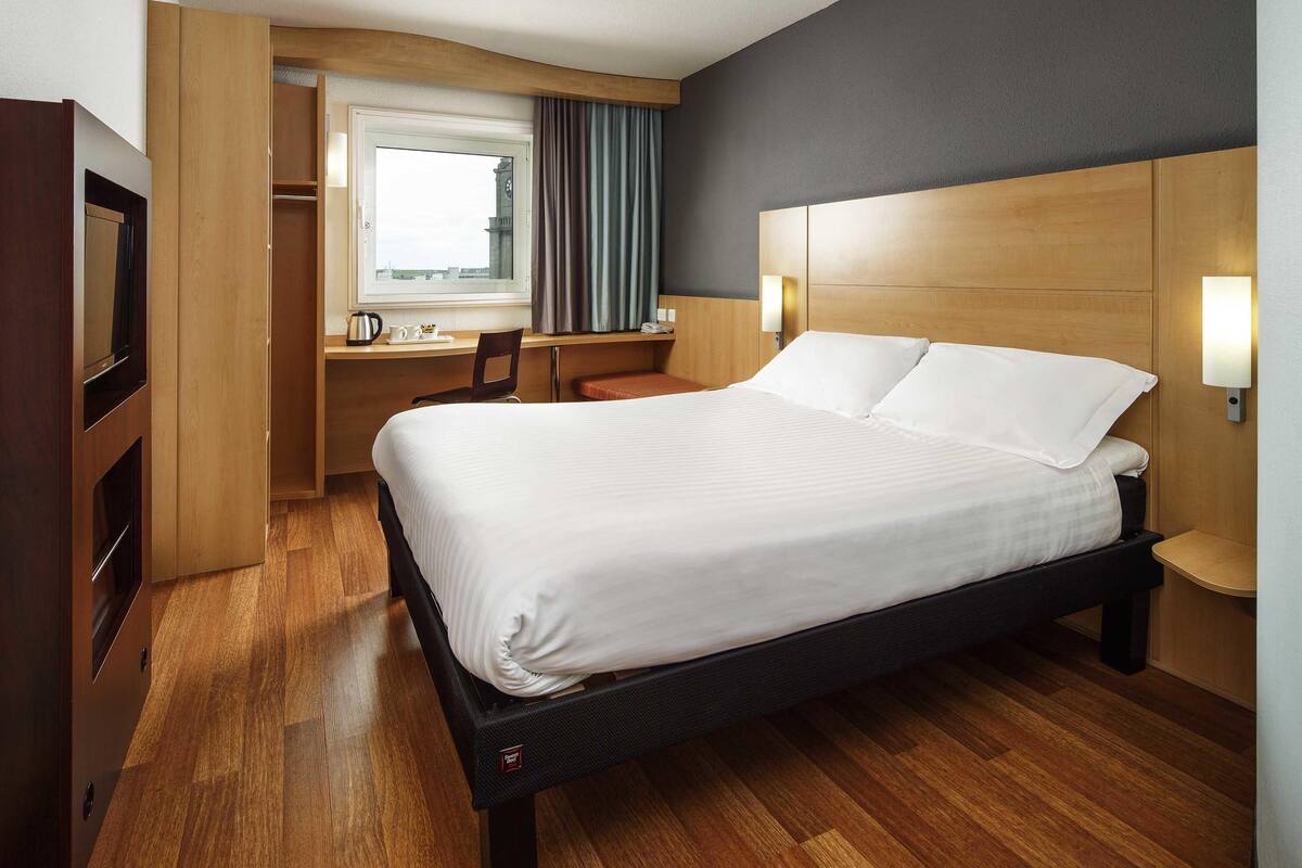 A hotel room with a bed, a desk, a chair, a window, and a clock.