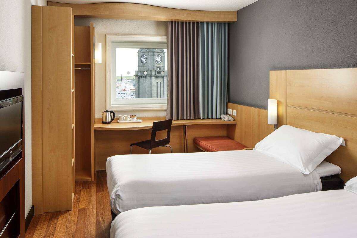 A hotel room with two beds, a desk, a chair, a TV, and a window with curtains.