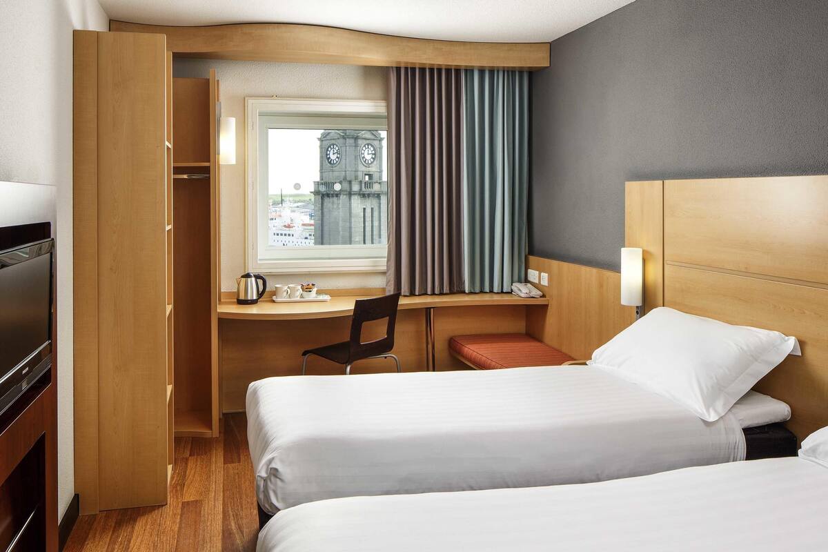 A hotel room with two beds, a desk, a chair, and a TV.