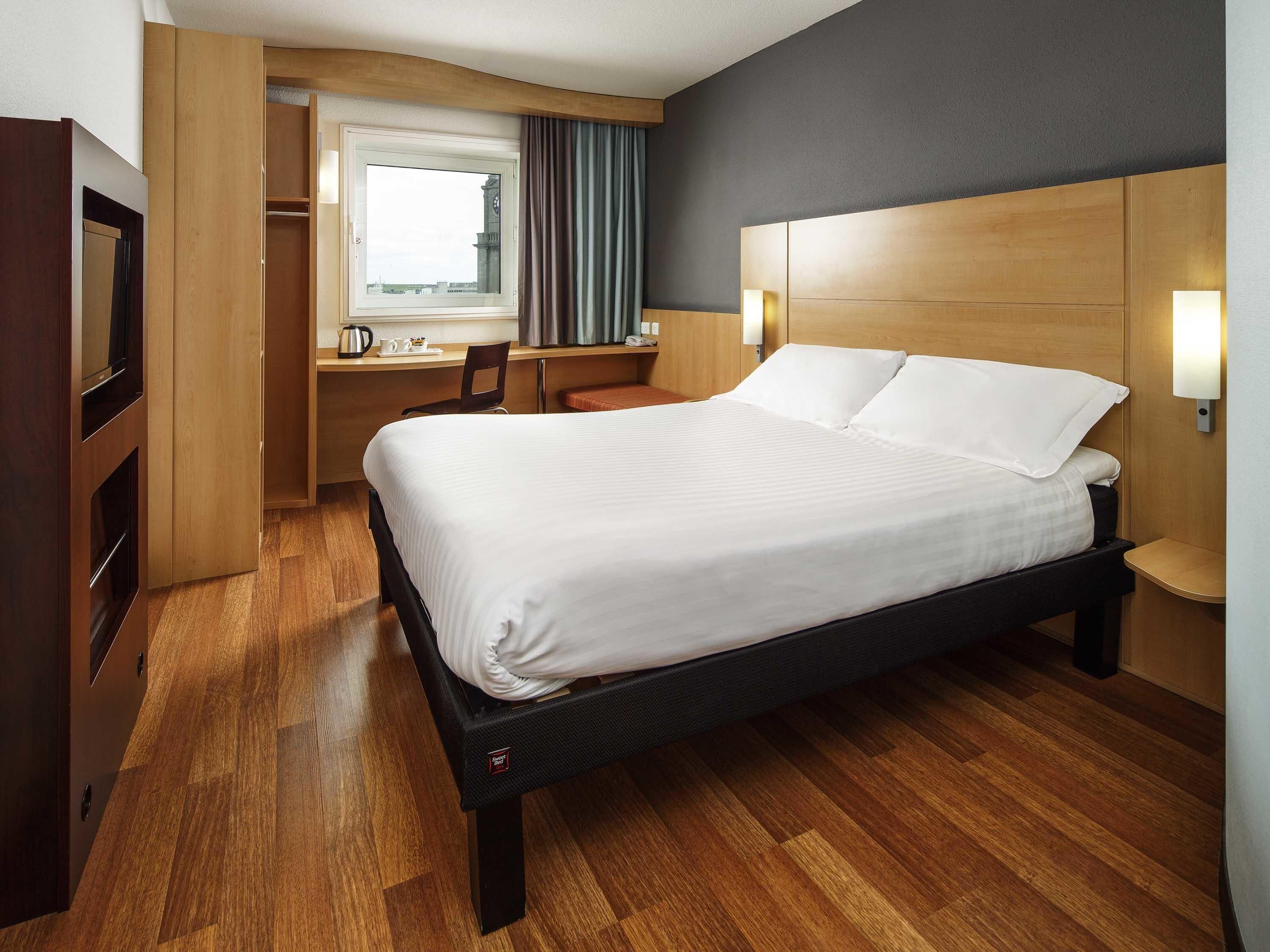 A hotel room with a bed, a desk, a chair, a window, and a clock.