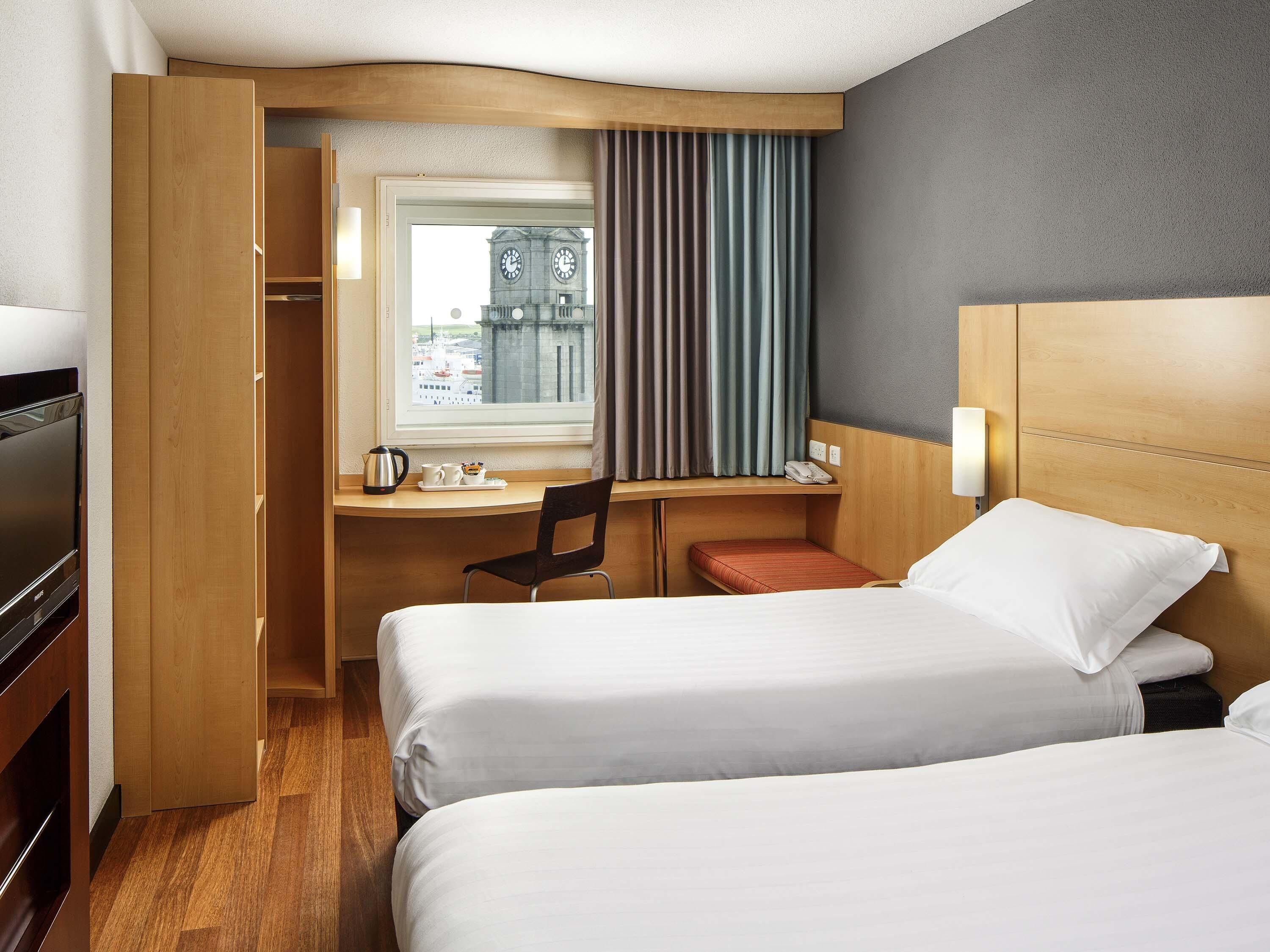 A hotel room with two beds, a desk, a chair, a TV, and a window with curtains.