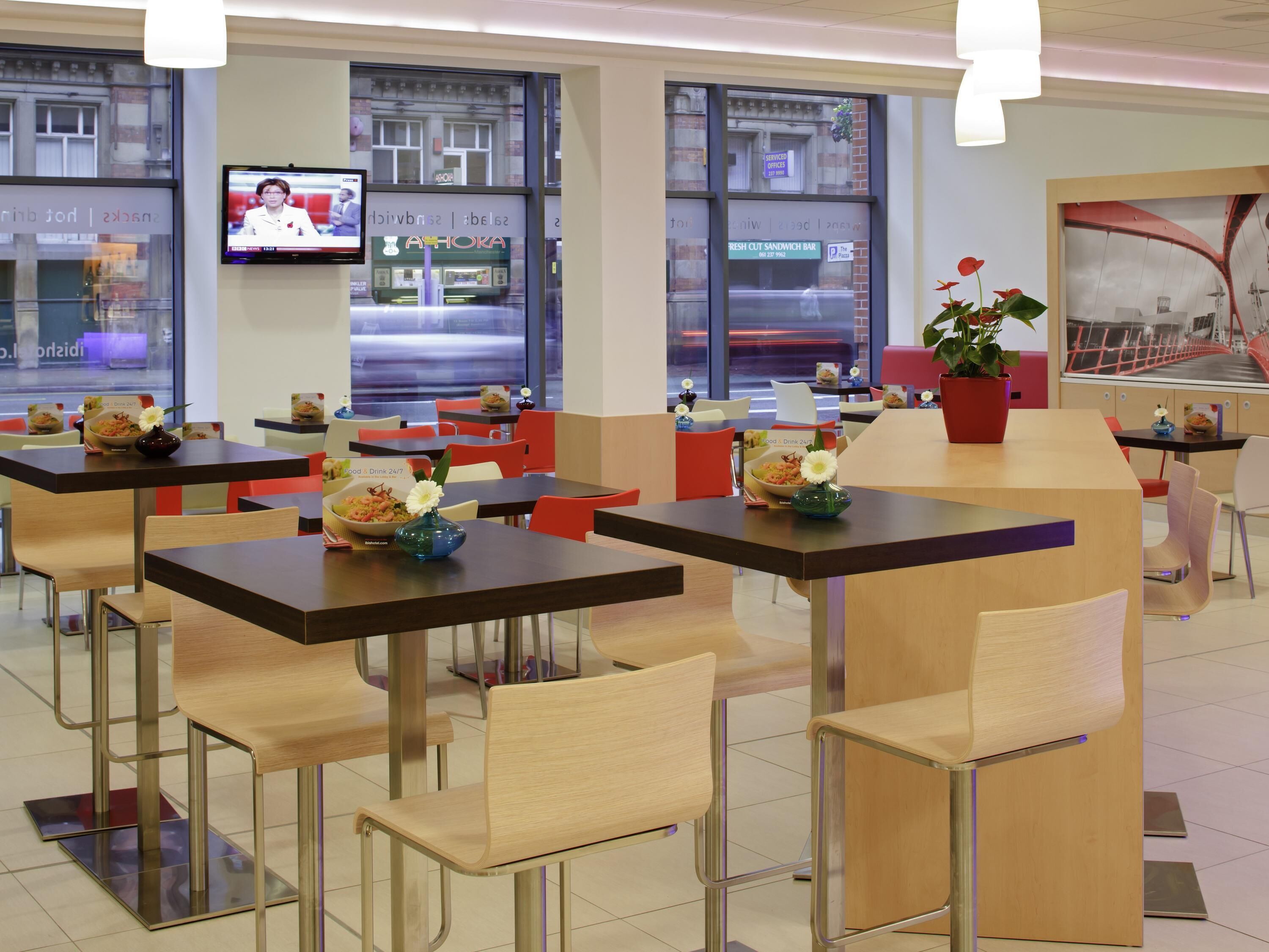 A modern restaurant with multiple tables and chairs, a television screen, and large windows.