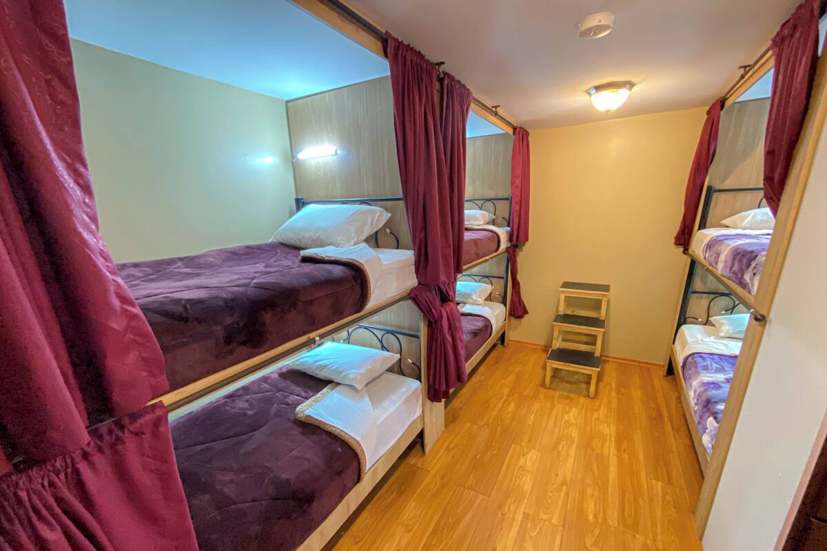 Habitación estrecha con literas, suelo de madera y una pequeña escalera de madera.