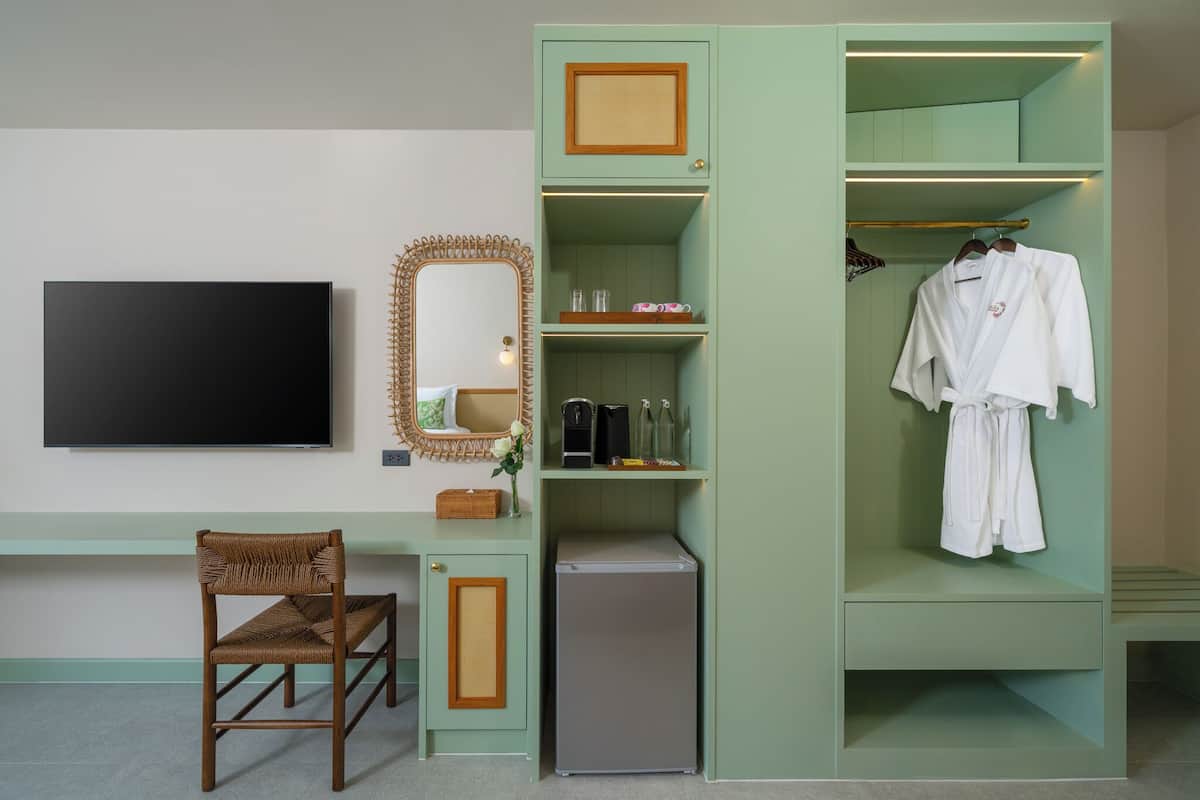 Une chambre dotée d’un téléviseur à écran plat, d’un fauteuil en osier, d’un petit réfrigérateur, d’un miroir et d’une armoire verte dans laquelle est suspendue une robe.