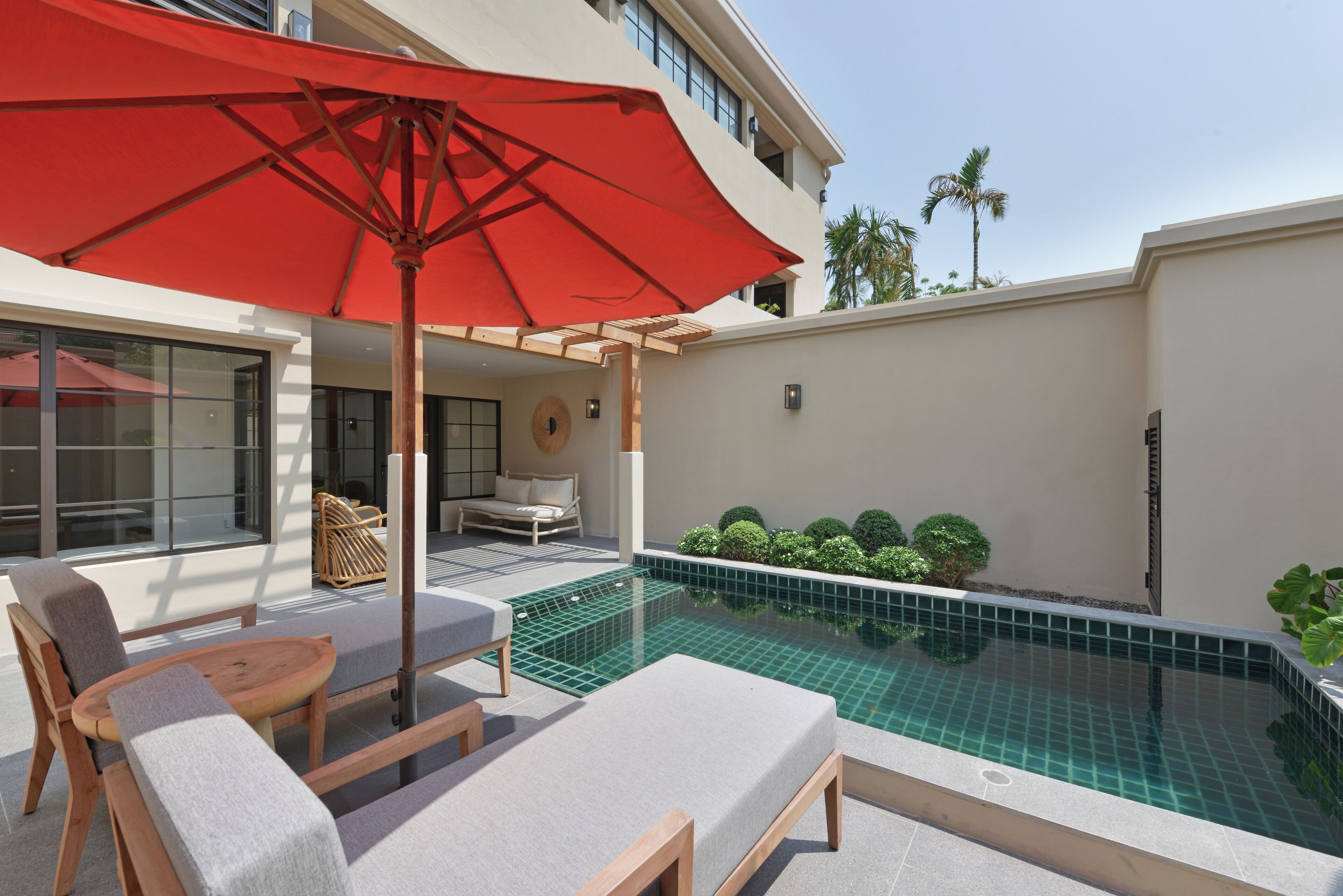 Un espace aménagé au bord de la piscine, avec des chaises longues et un parasol rouge, une petite piscine et un bâtiment doté d’un balcon.