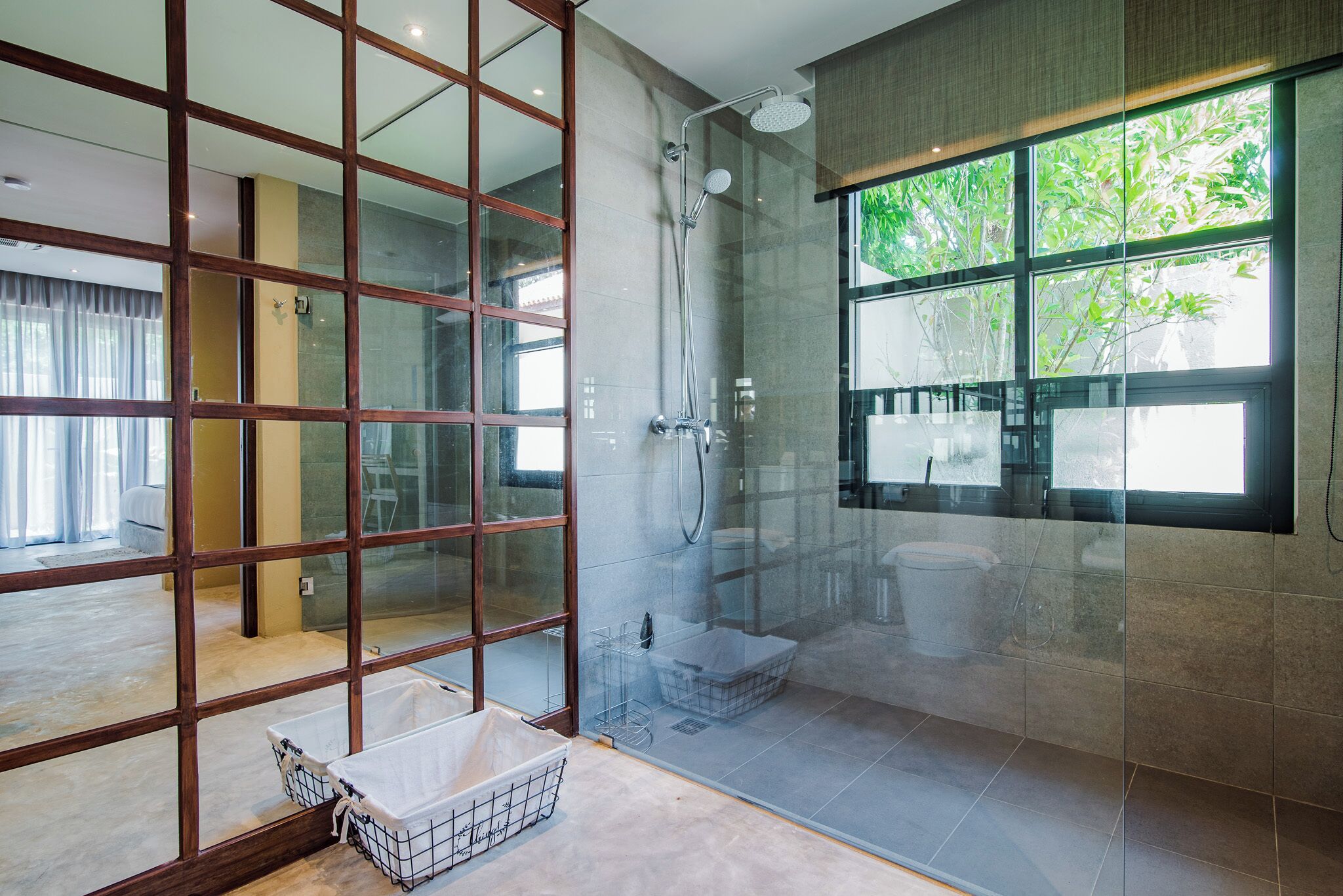 A modern bathroom with a glass shower enclosure, a large window with a view of greenery, and a wooden partition.