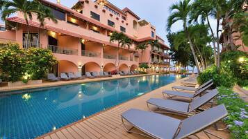 A poolside area with lounge chairs, a swimming pool, and a hotel building with balconies and palm trees.