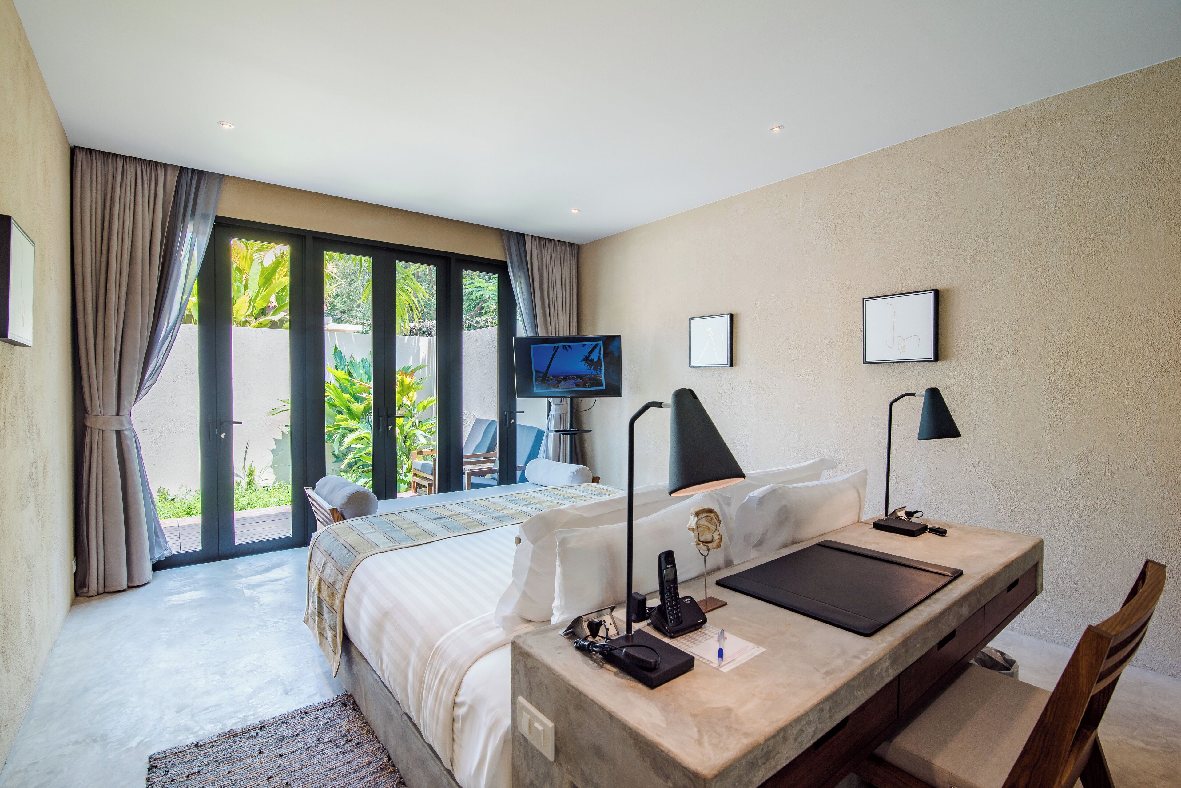 A modern hotel room with a large bed, a desk with a chair, a television, and a view of greenery outside.