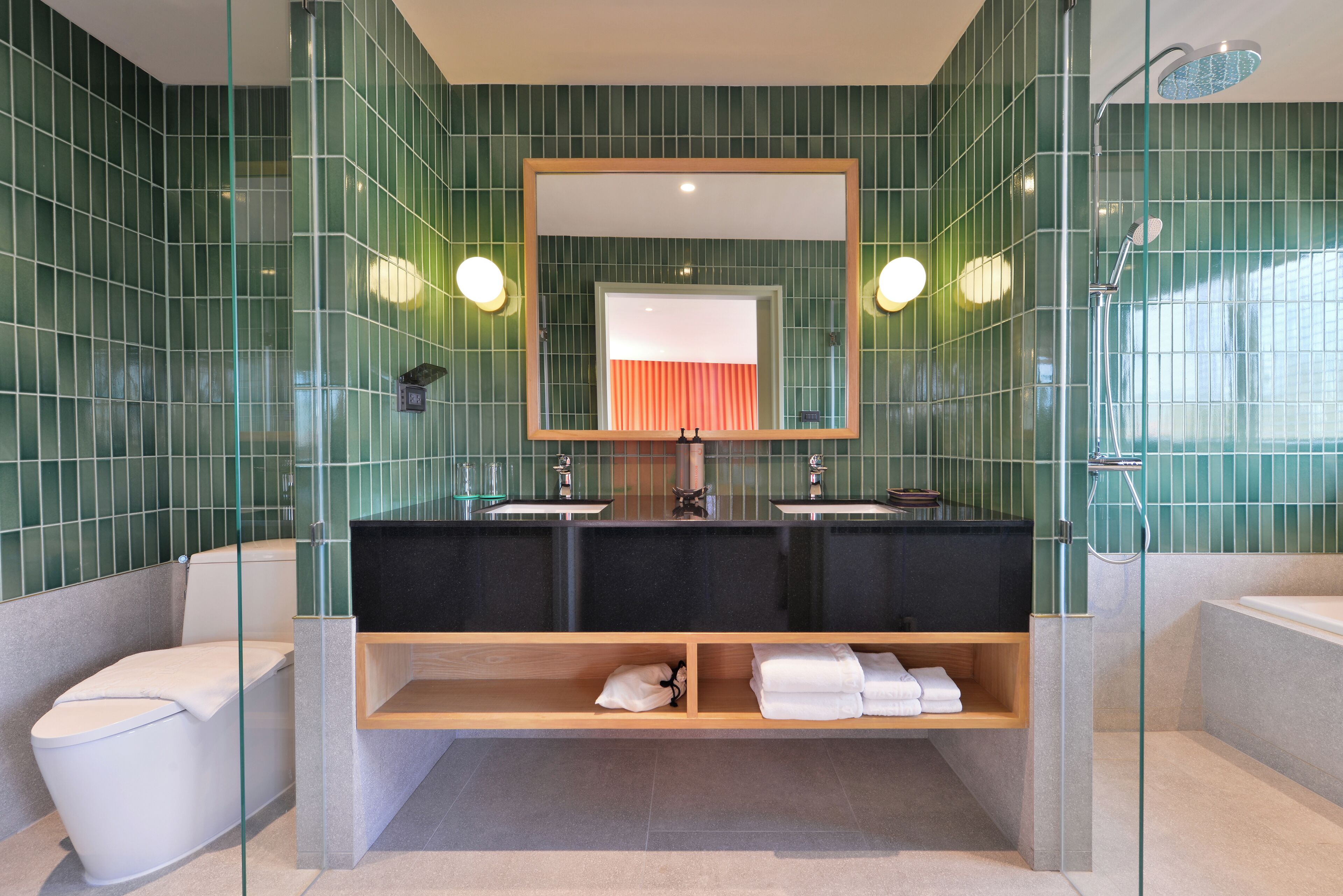 A modern bathroom with green tiled walls, a dual sink vanity, a toilet, and a glass-enclosed shower area.