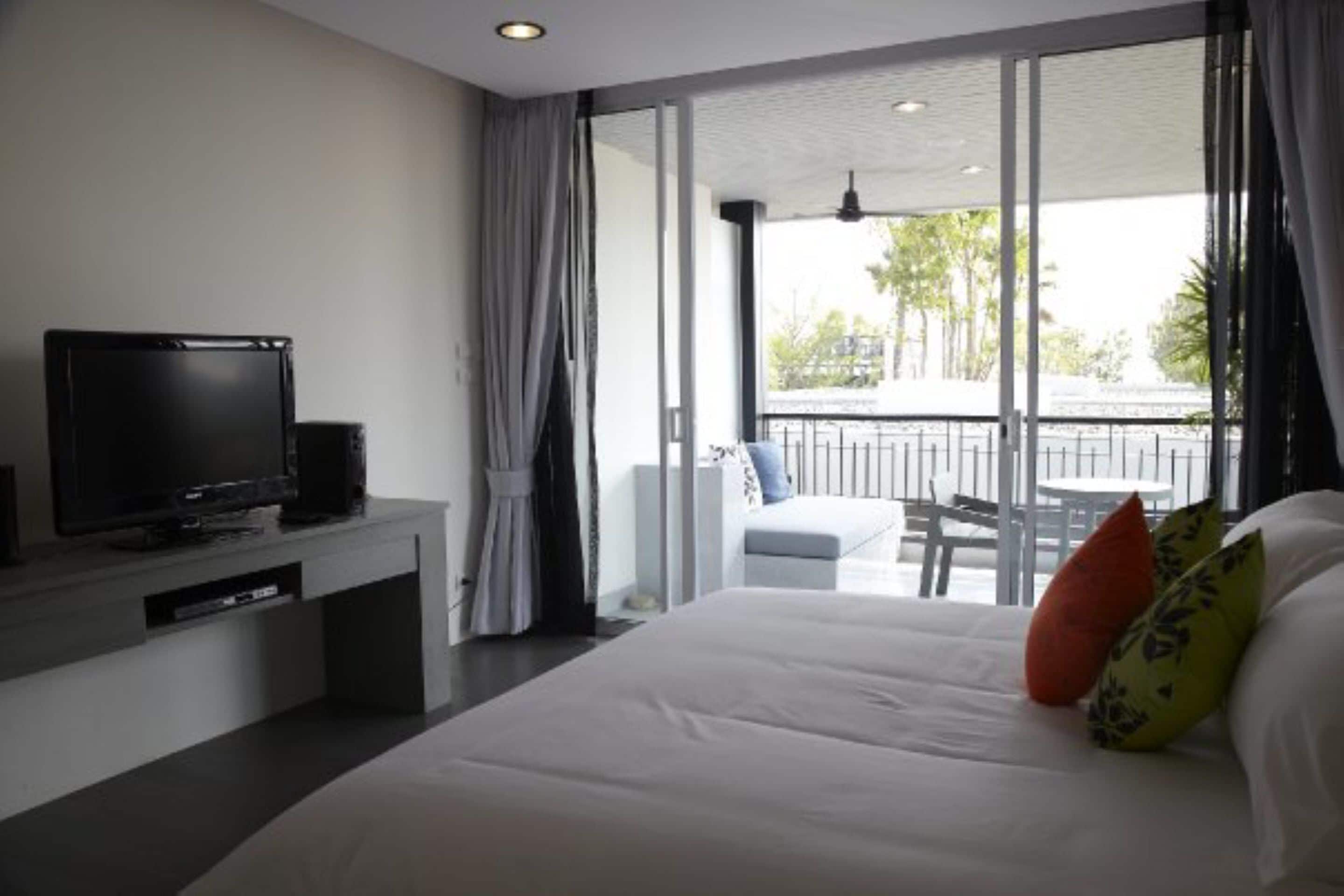 A modern hotel room with a large bed, a TV on a stand, a balcony with a view of palm trees, and a dining area with a table and chairs.