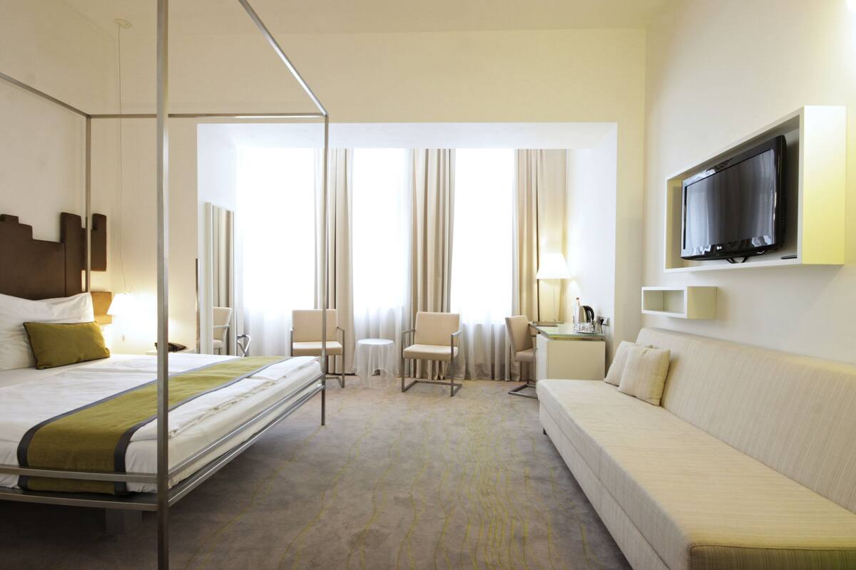 A hotel room with a four-poster bed, a sofa, a TV mounted on the wall, and a desk with a lamp.