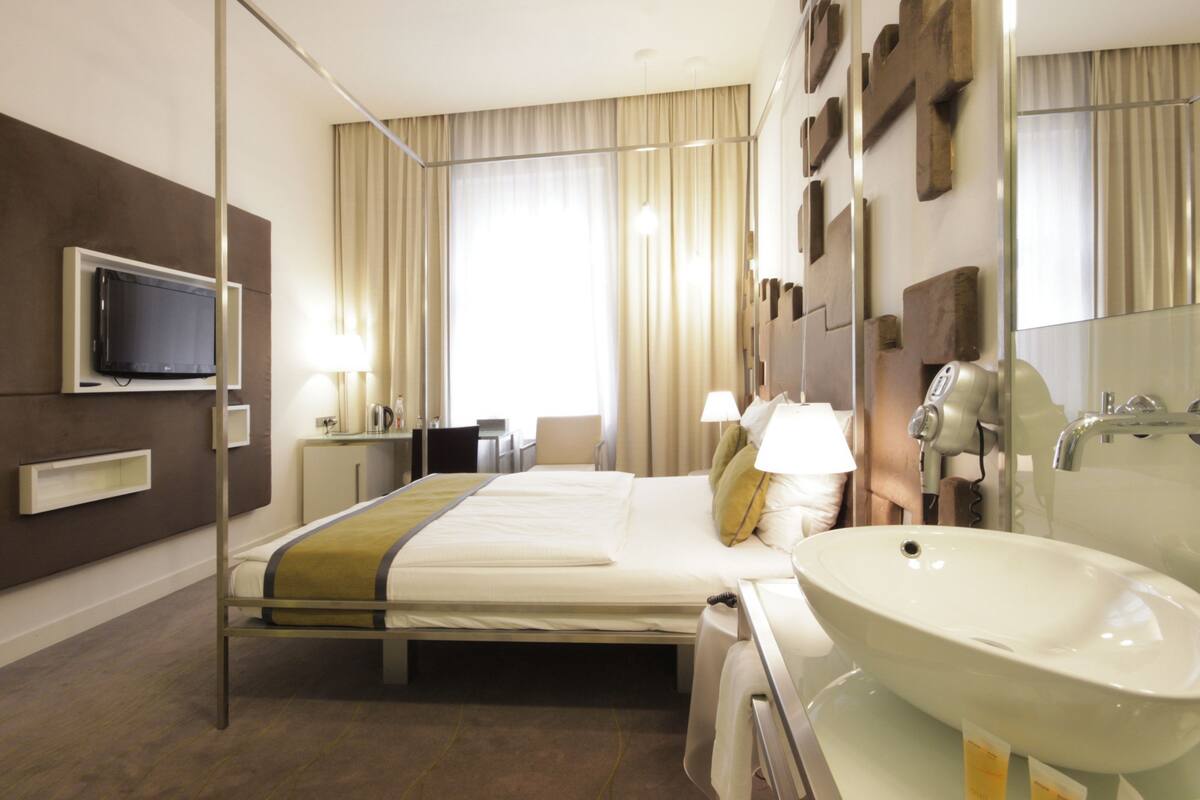 A hotel room with a bed, bedside table, lamp, TV, and a large mirror.