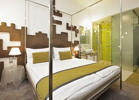 Quarto de hotel moderno com uma cama grande, mesas de cabeceira, toucador com espelho e chuveiro em cabine de vidro.