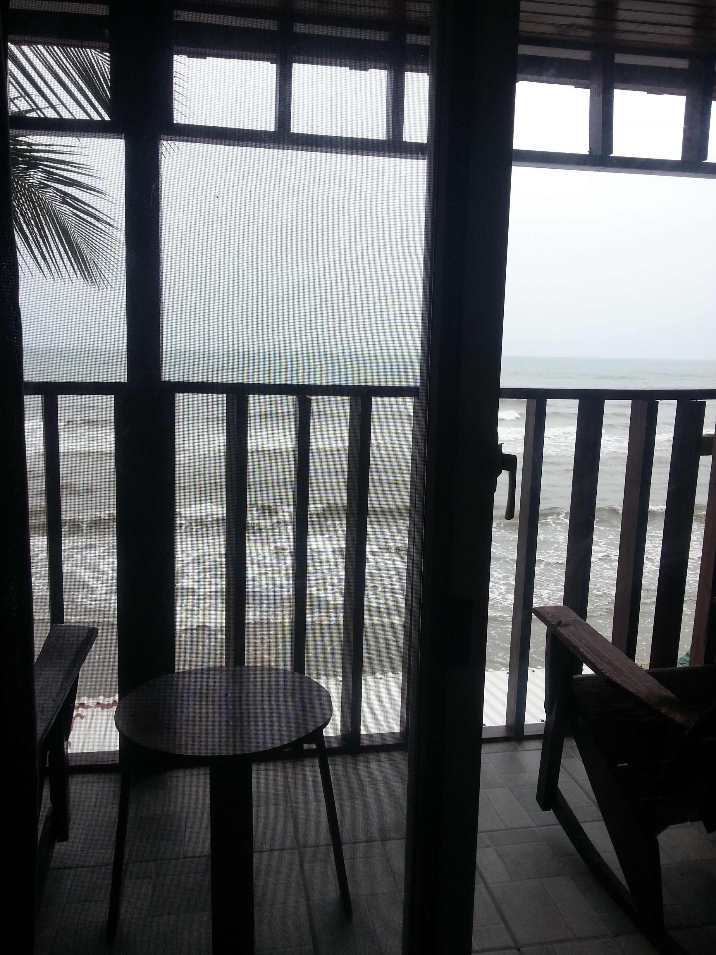 A view from a covered balcony overlooking a beach with waves. There are two wooden chairs and a small round table.