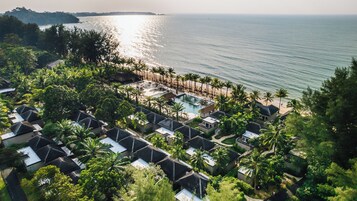 Vista aérea de un resort con varias cabañas, un área de piscina central y una playa con palmeras.