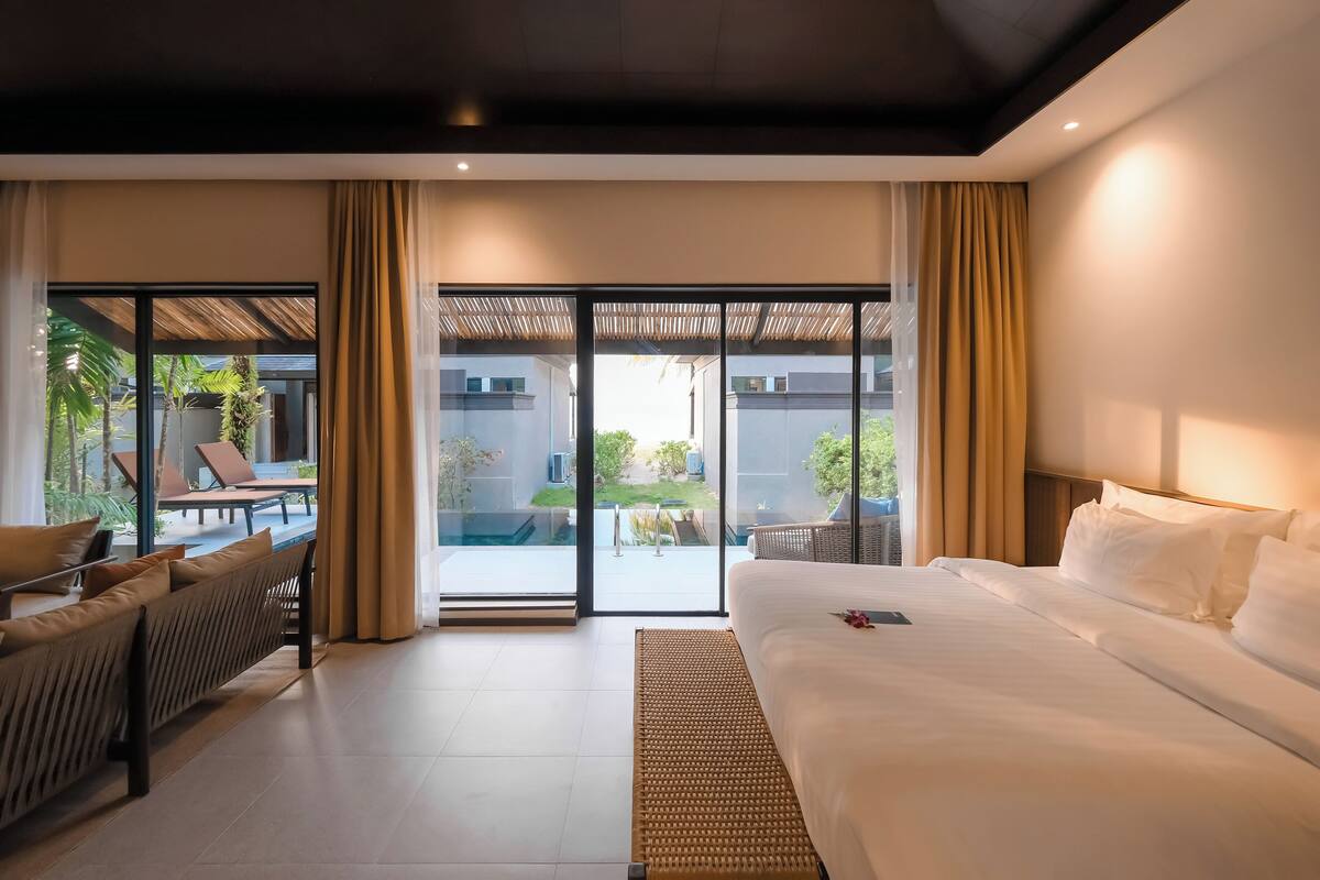 Una habitación de hotel moderna con una cama grande, un sofá y un balcón con vistas a la piscina y la vegetación.
