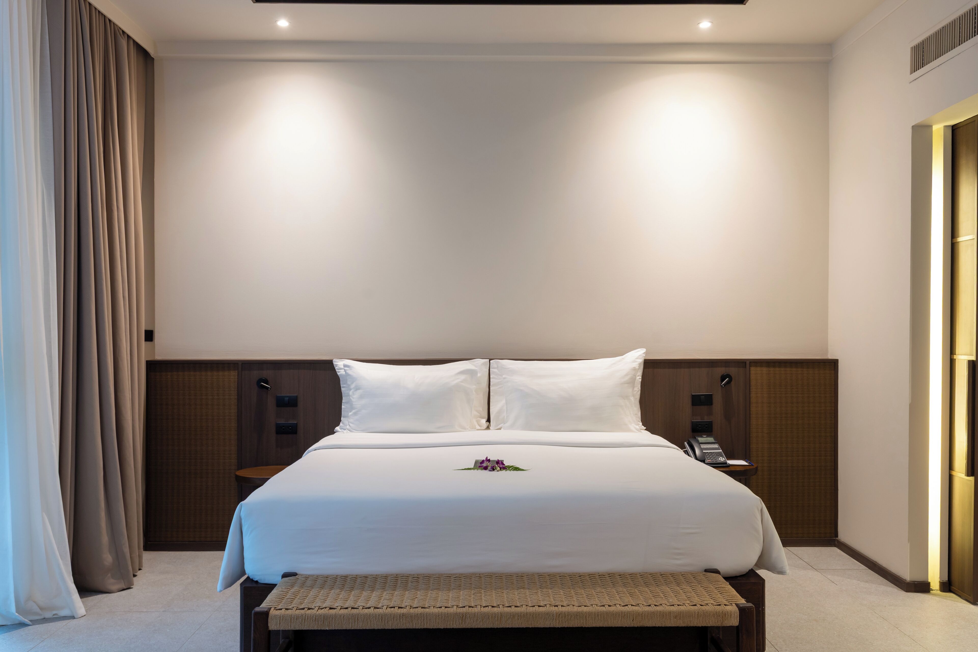 A modern hotel room with a large bed, a bedside table, a lamp, and a bench.