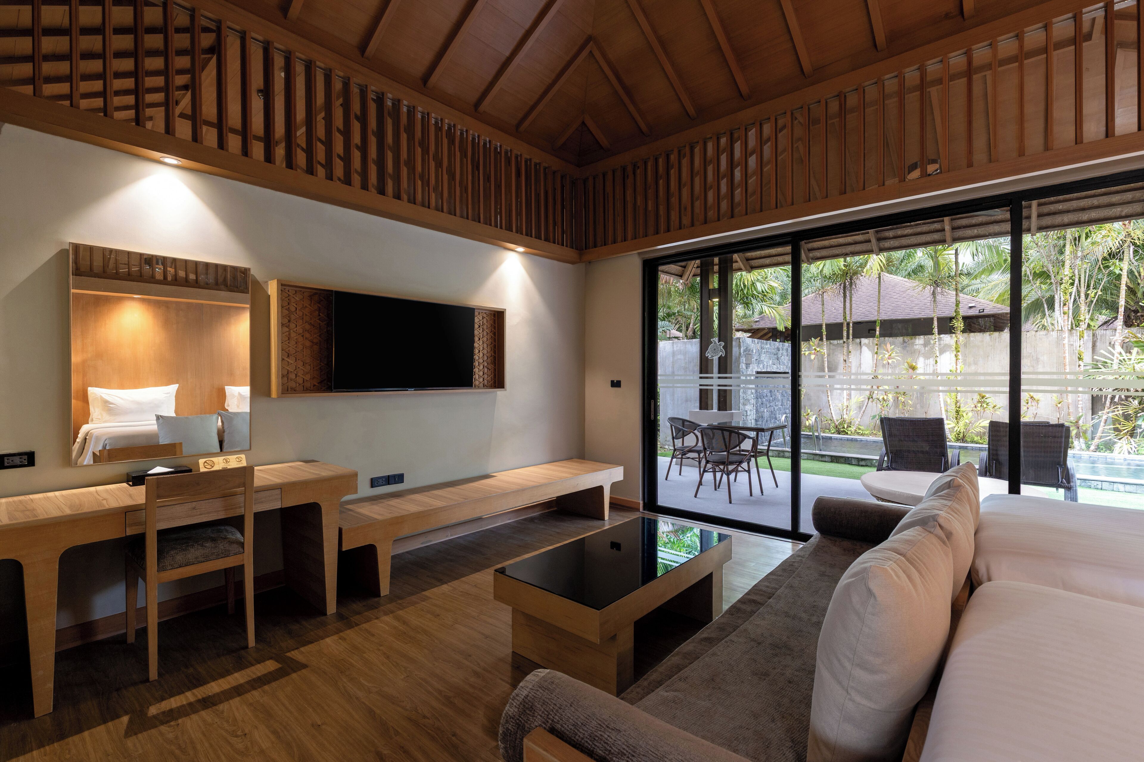 Una habitación moderna con escritorio de madera, sofá y un amplio ventanal con vistas a una piscina y palmeras.
